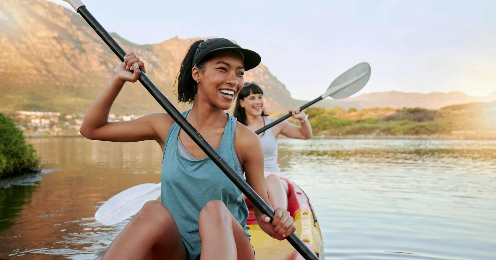 Two women kayaking on a calm river with scenic mountains in the background.
